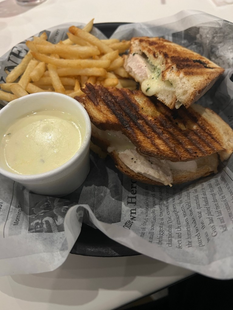 Grilled pieces of bread with chicken and pesto pieces, next to a cup of white alfredo sauce and in the back, a side of yellow french fries.